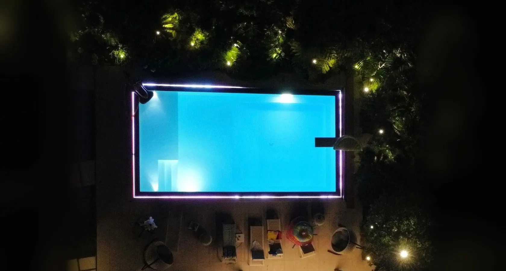 Piscina illuminata con luci LED vista dall&rsquo;alto di notte, tra le piante di un giardino privato